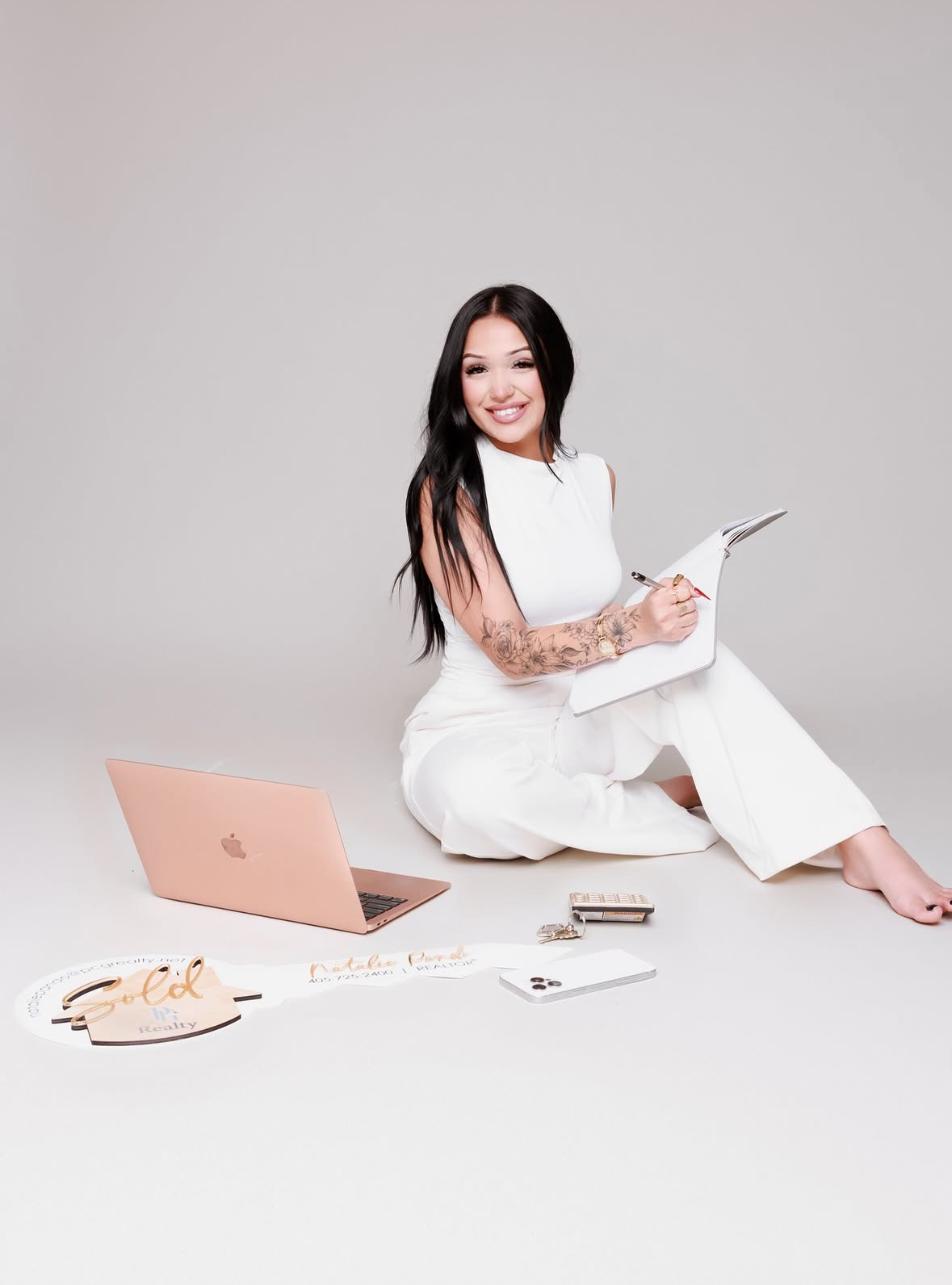 Woman in white outfit sitting on floor with rose gold MacBook, notepad, and branded accessories in professional photo.