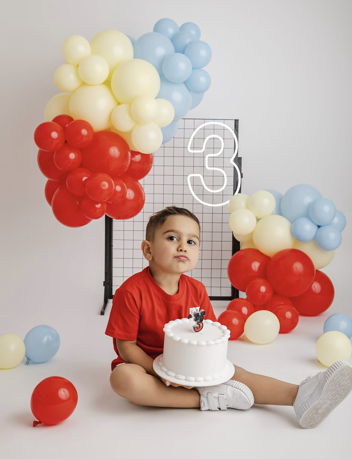 A toddler in a red shirt holds a white birthday cake, surrounded by red, blue, and yellow balloons near a number 3 sign.