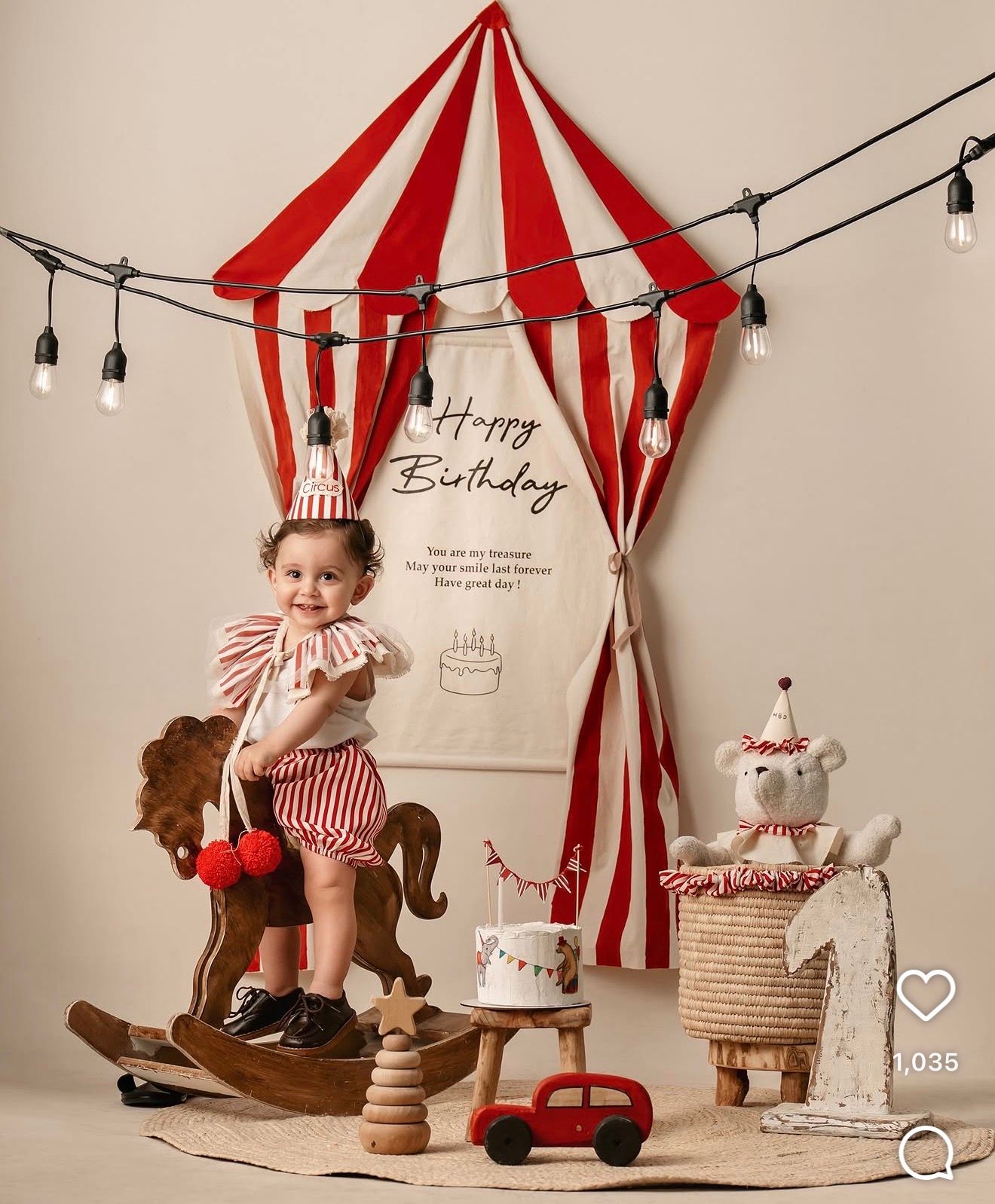 Toddler in circus outfit sits on rocking horse beside birthday cake and tent backdrop for first birthday photo.