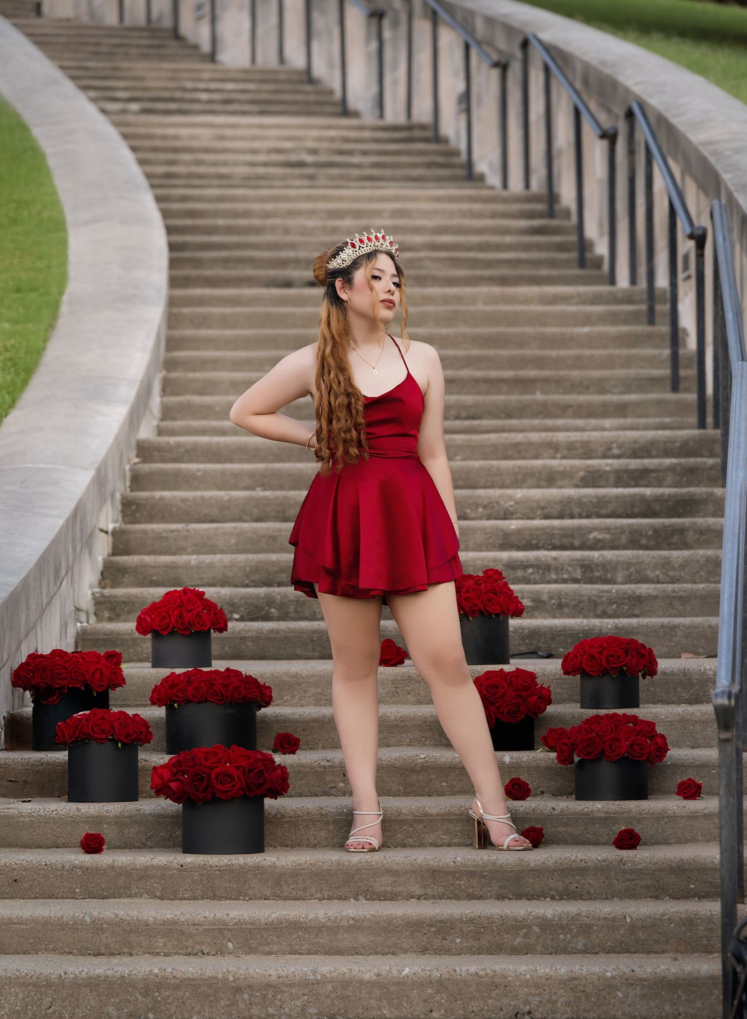 Young woman in red mini dress and crown poses on stone steps surrounded by black boxes of red roses.