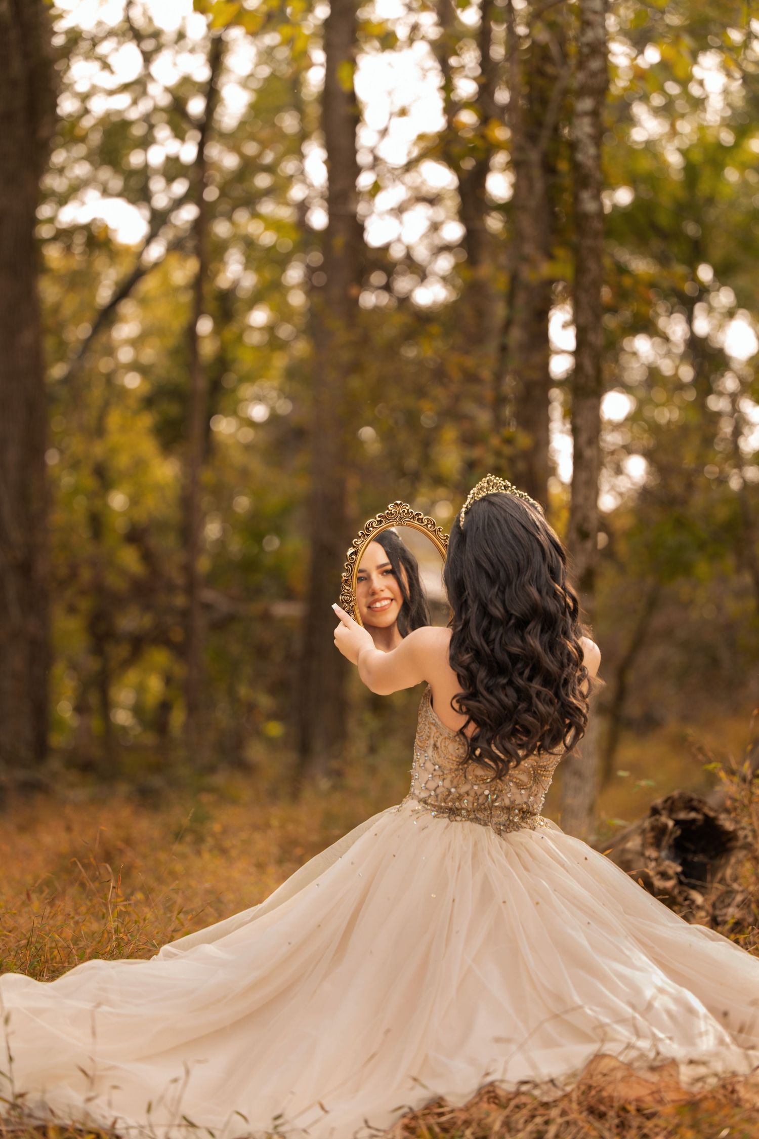 A woman in a beige ball gown and gold crown holds a mirror in an autumn forest, showing her smiling reflection.