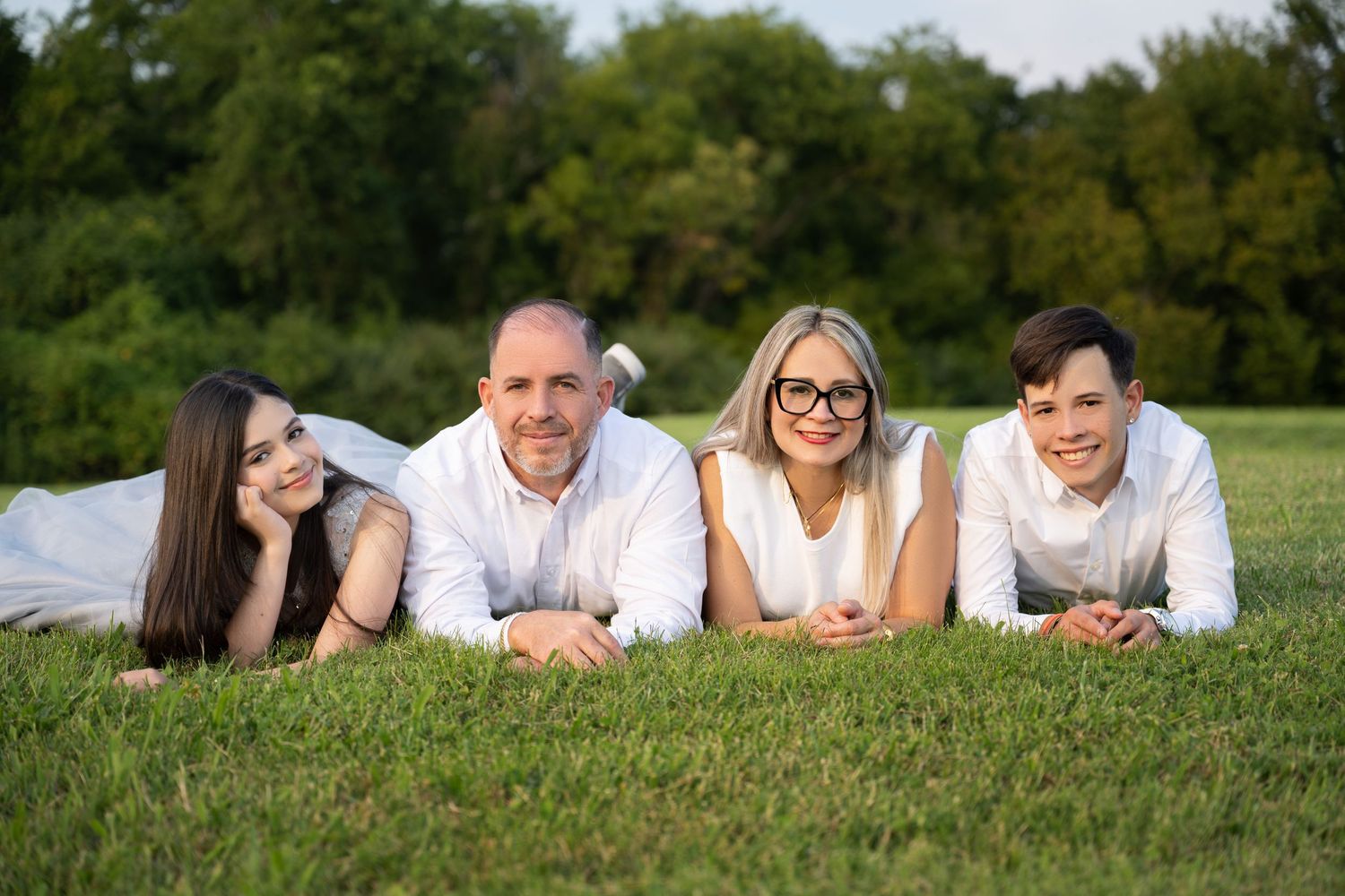 Family of four lying on green grass smiling, wearing white outfits, with trees in the background.