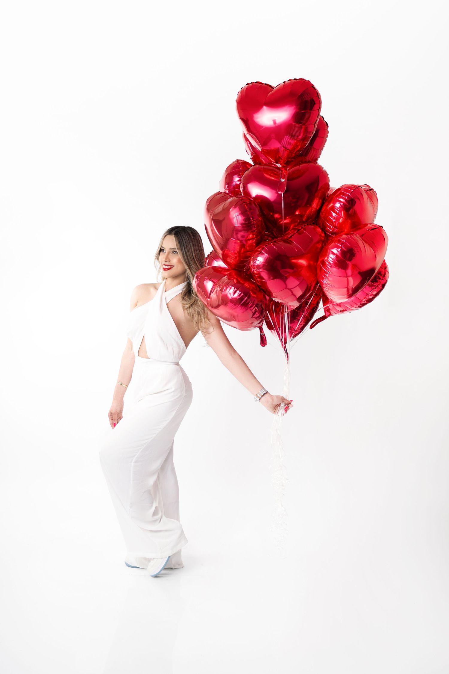 Woman in a white halter jumpsuit holds a bunch of red heart-shaped foil balloons against a white background.