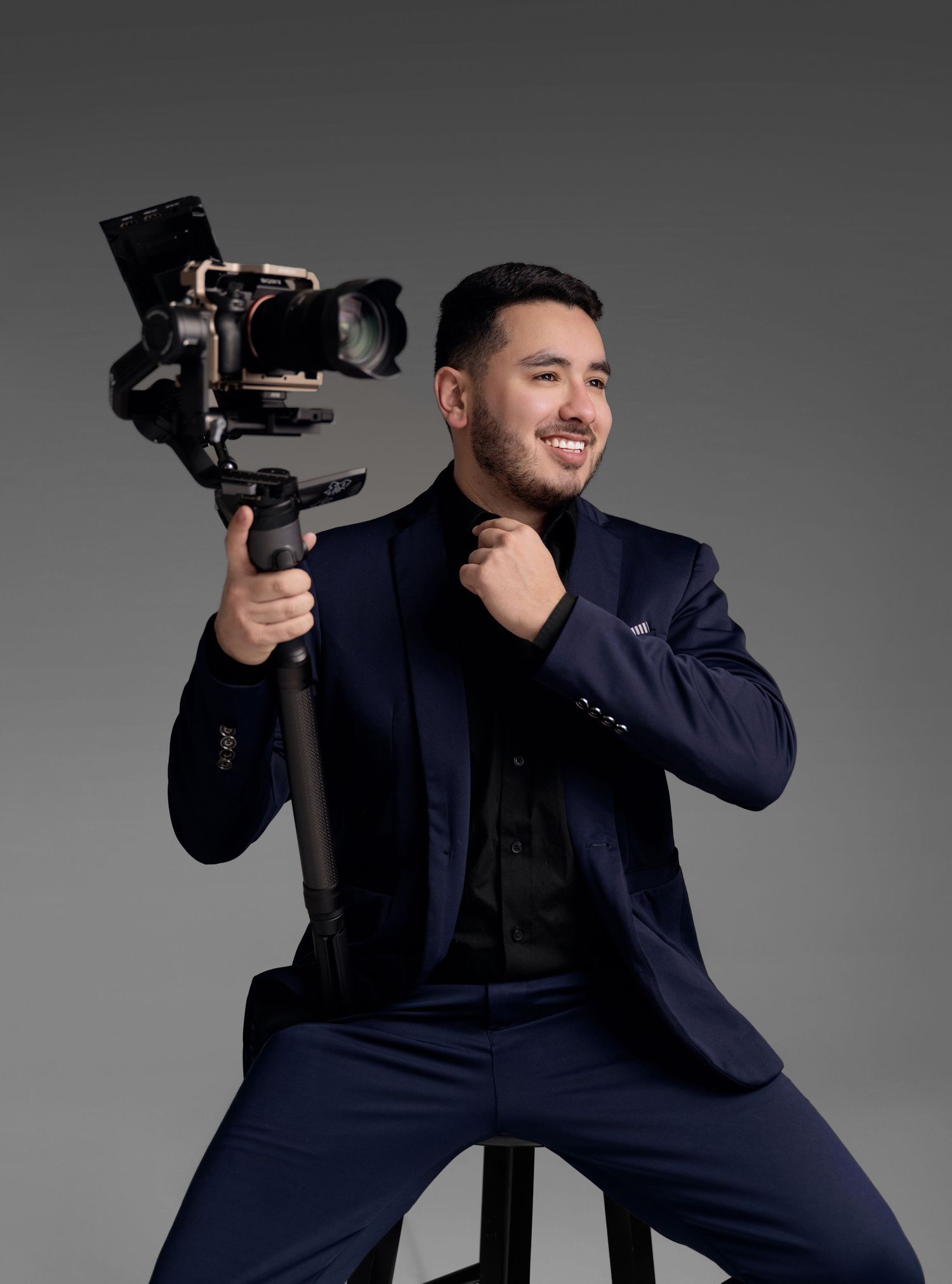 Smiling videographer in a navy suit sits on a stool holding a professional camera on a gimbal stabilizer against a gray studio backdrop.