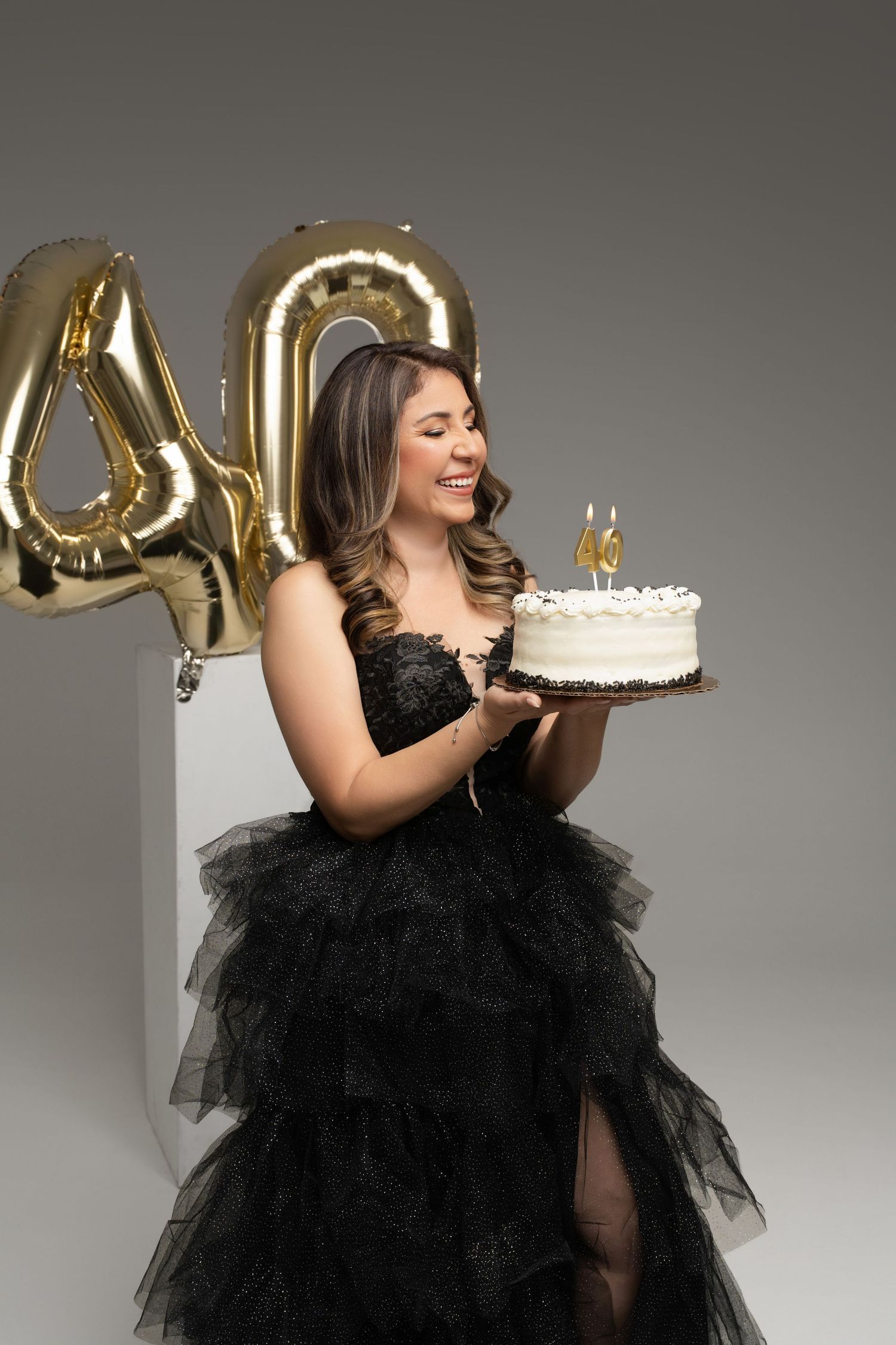 Woman in black gown smiling at 40th birthday cake with gold number balloons in background.