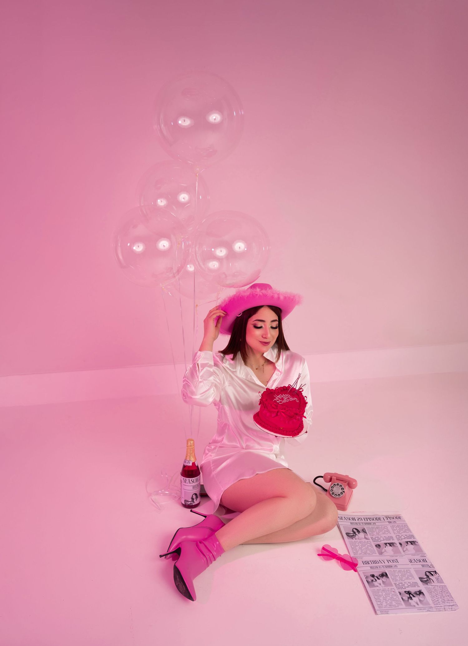 Woman in pink cowboy hat and boots sits on floor with balloons, champagne, and heart purse in pink studio.