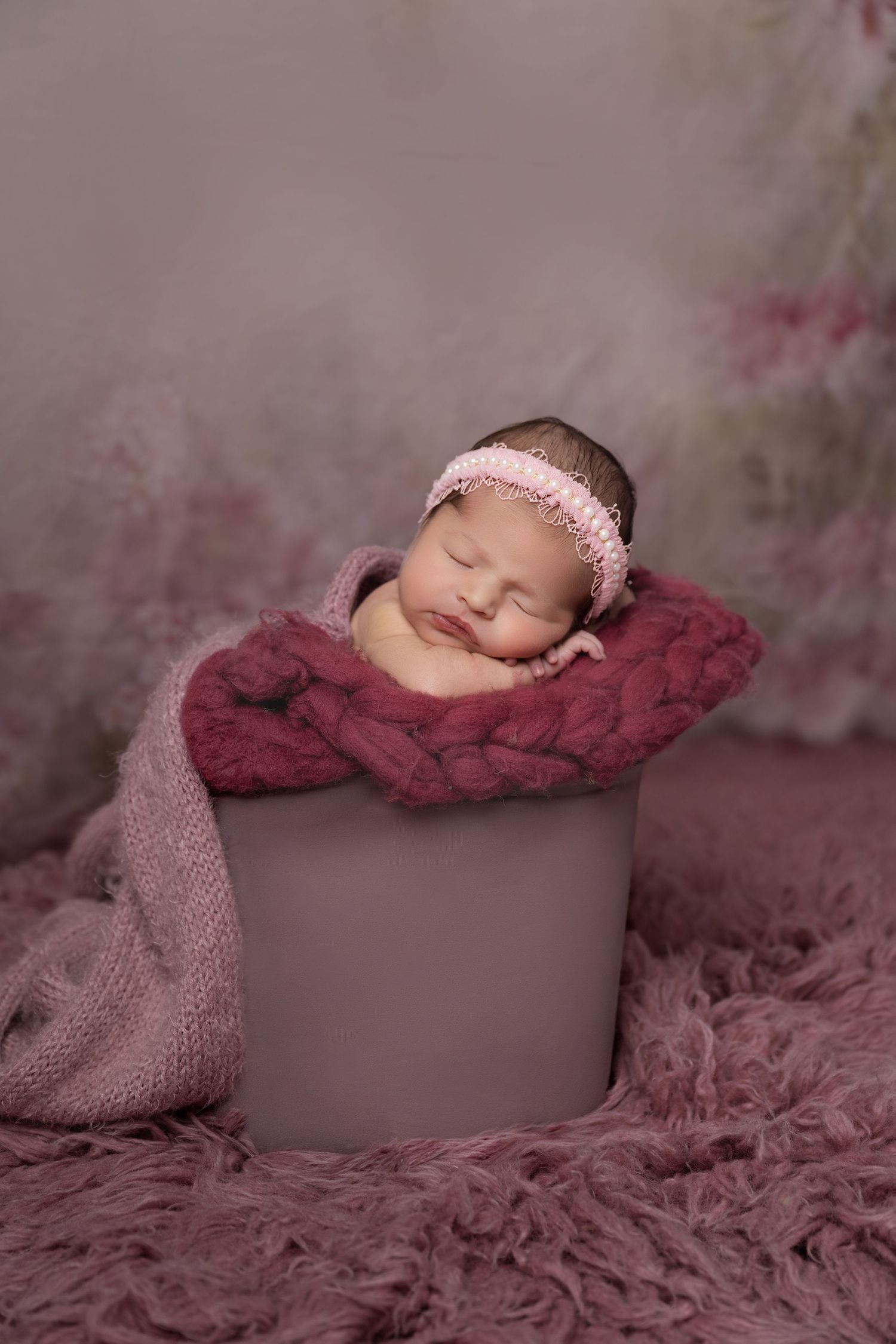 Sleeping newborn baby in a pink bucket with a knit blanket and headband on a floral backdrop.