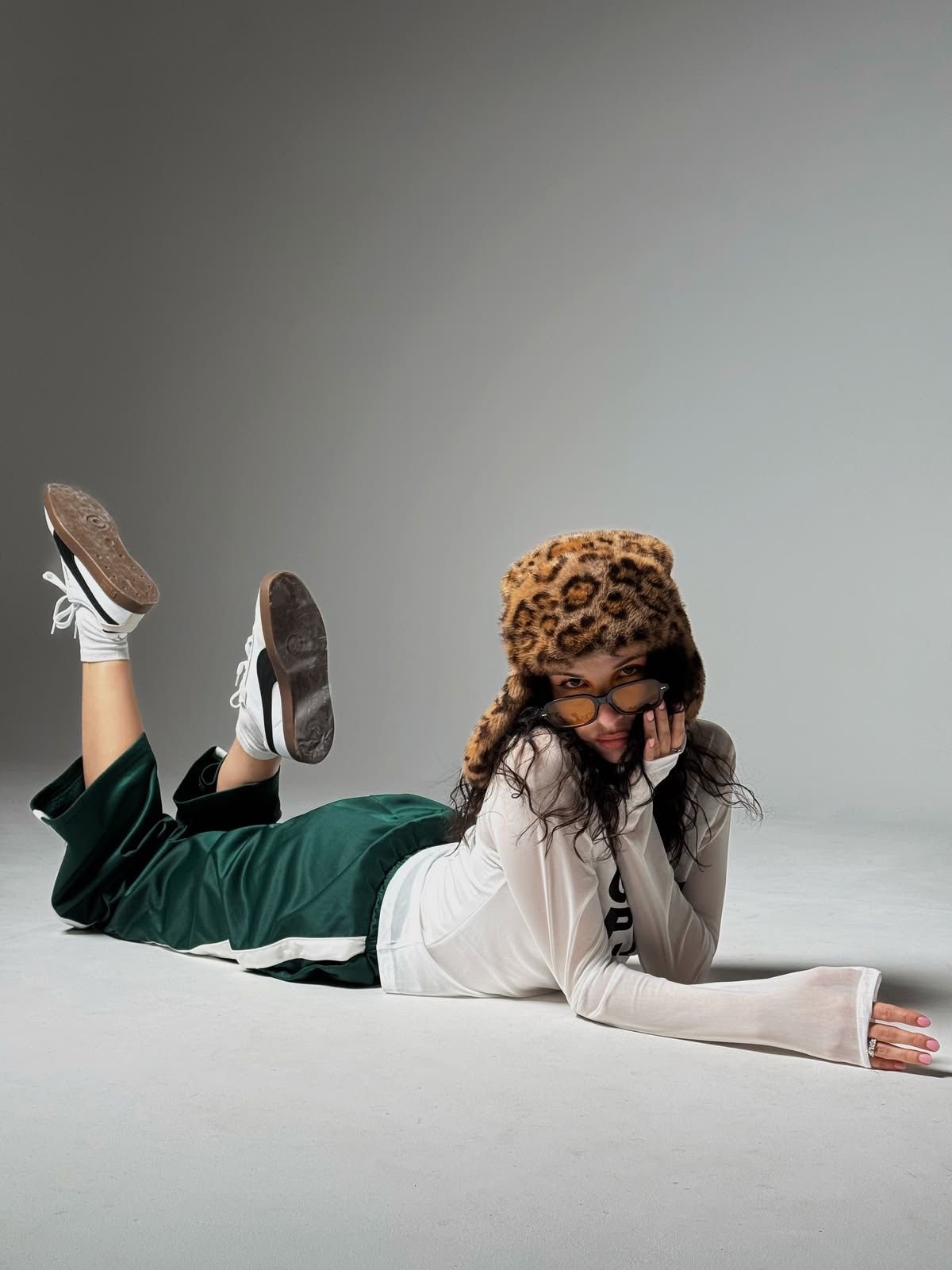 Person lying on floor wearing leopard bucket hat, green pants, white top, and sneakers in a studio setting.
