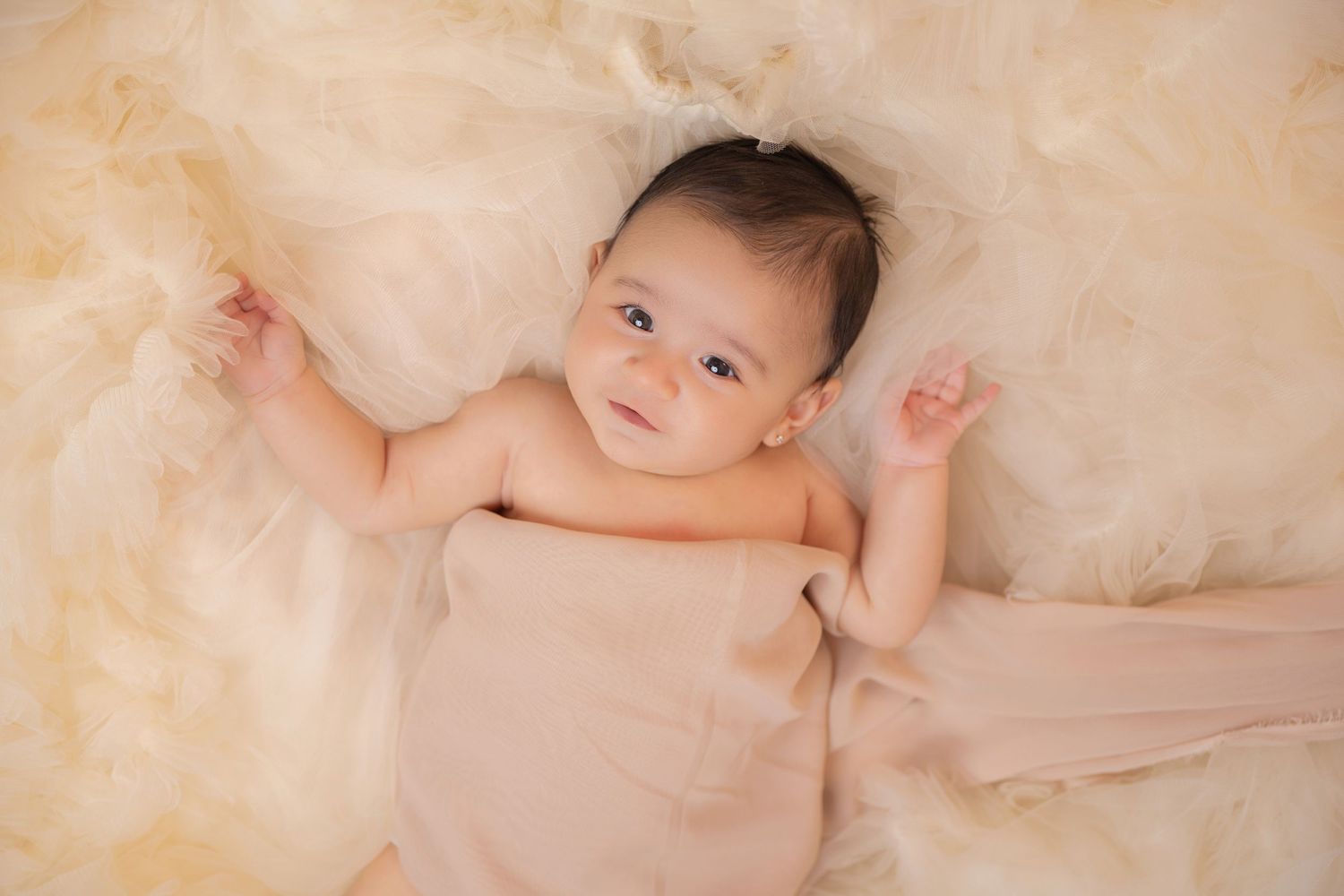 Newborn baby wrapped in a soft pink blanket, lying on cream tulle, smiling with bright eyes in a studio photo.