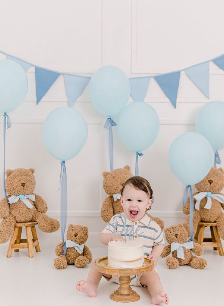 First birthday cake smash photoshoot setup with teddy bears, blue balloons, and bunting decorations in soft pastel colors.