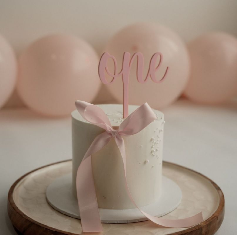 A white frosted cake with pink ribbon sits on a rustic wooden cake stand against a soft pink background.