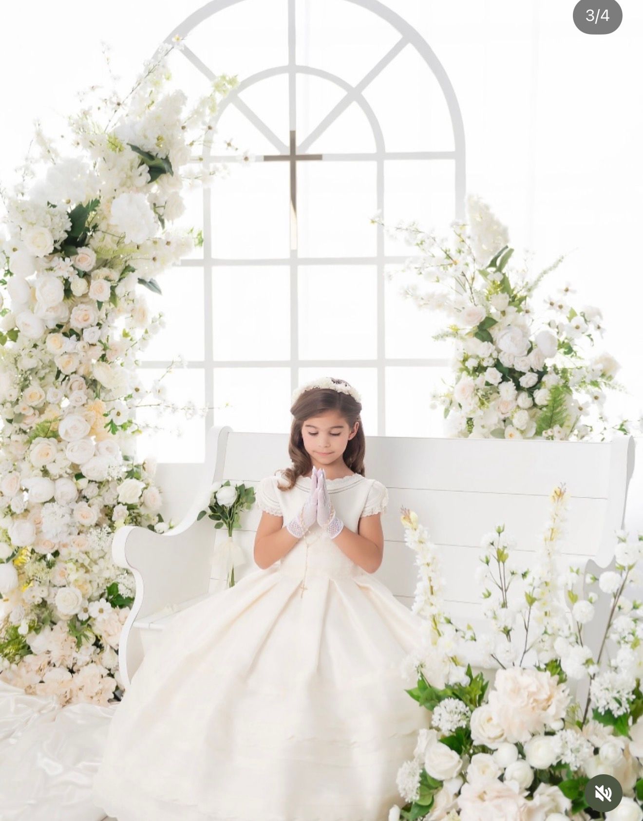 Young girl in white First Communion dress praying hands, surrounded by white floral arch with cross backdrop.