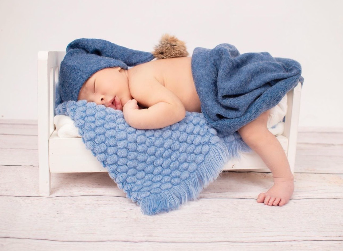 Sleeping baby wearing blue outfit lying on white bedding with knitted blue blanket.