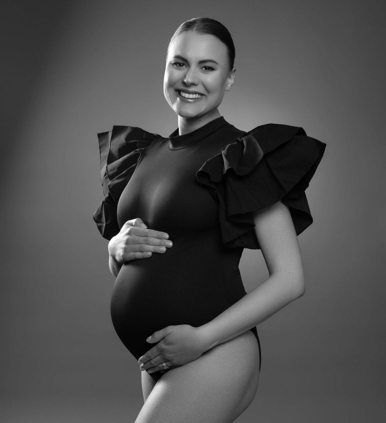 Black and white maternity photo of a smiling pregnant woman in a ruffled black bodysuit cradling her baby bump.