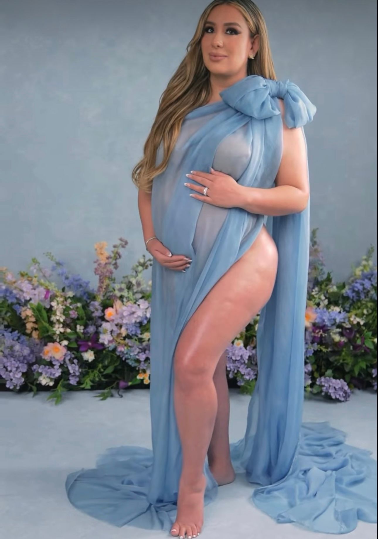 Pregnant woman in elegant blue chiffon maternity gown with bow detail, posing before floral backdrop.