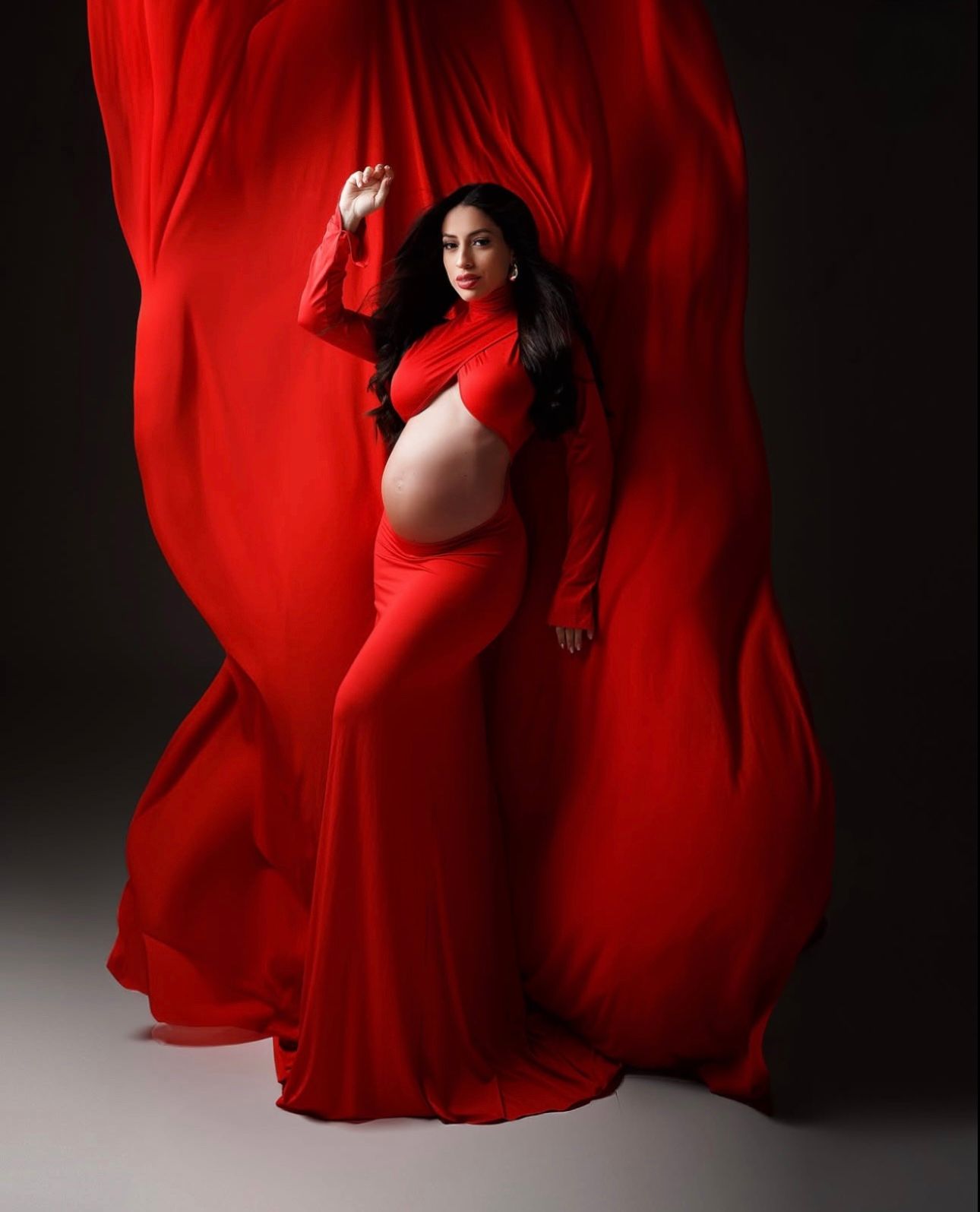 Pregnant woman in flowing red gown with billowing fabric, maternity photography studio portrait.