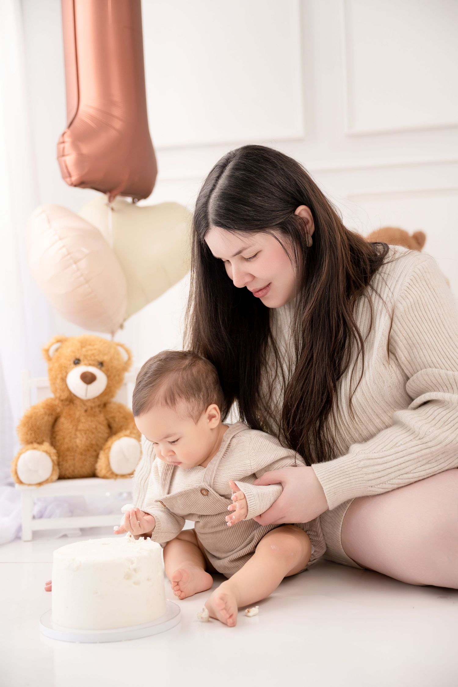 Mother and baby celebrating first birthday with white cake, teddy bear, and rose gold balloons in bright white room.