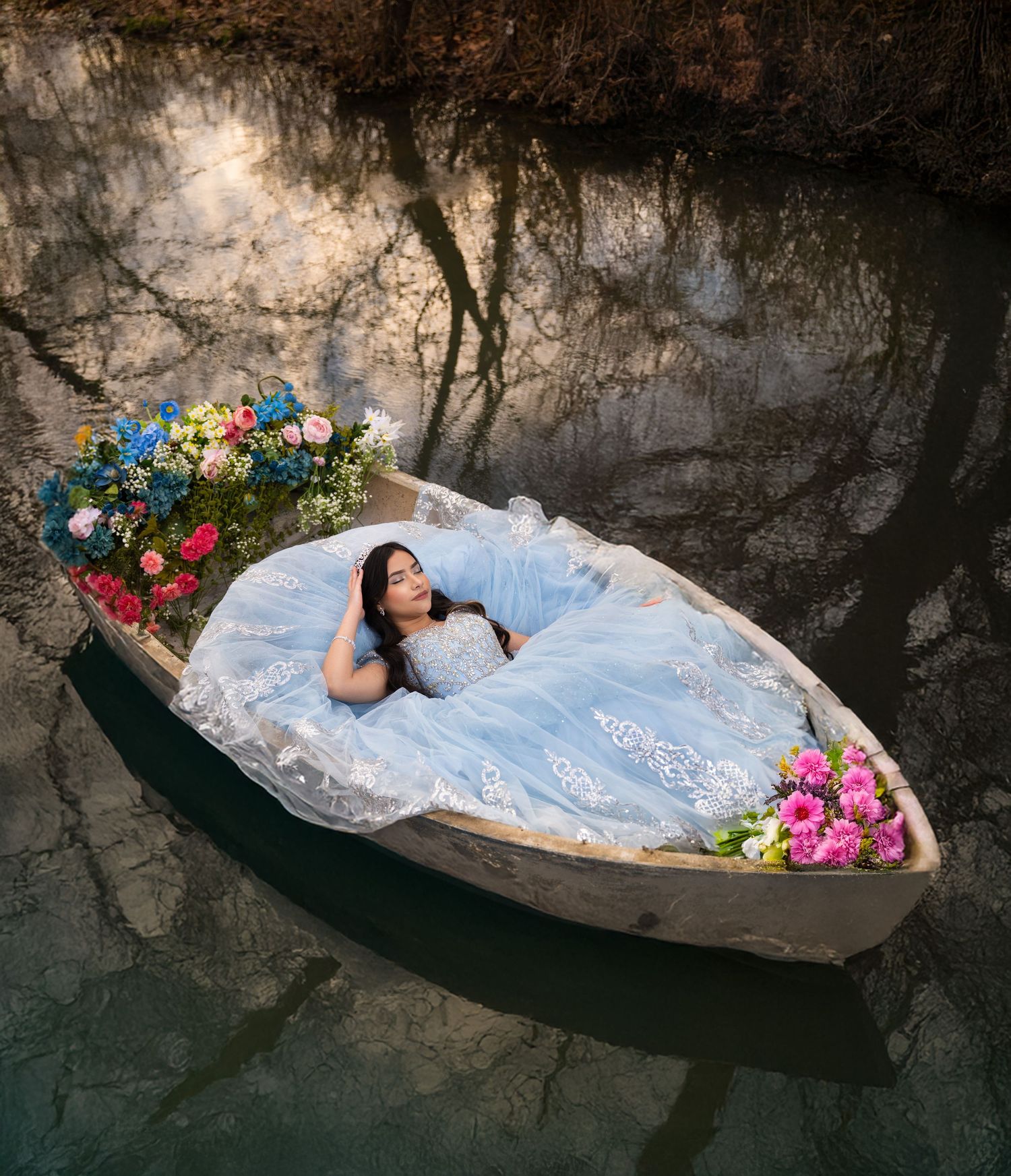 A woman in a blue lace gown reclines in a flower-adorned wooden boat on a calm reflective waterway.