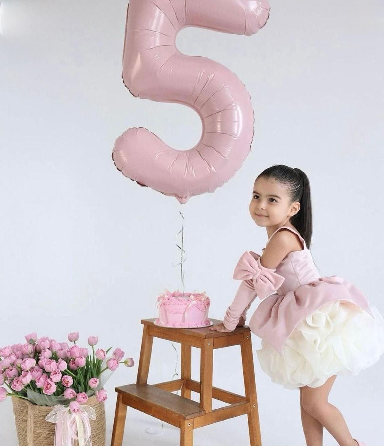 A young girl in a pink dress celebrates her 5th birthday with a cake and a large pink number 5 balloon.