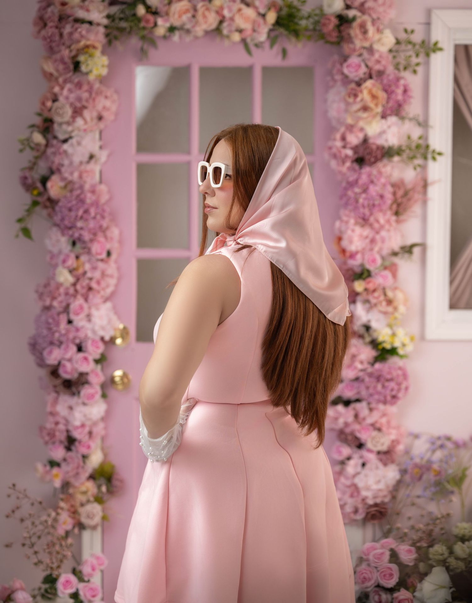 Woman in pink dress and white sunglasses with pink silk headscarf poses before floral-framed mirror.