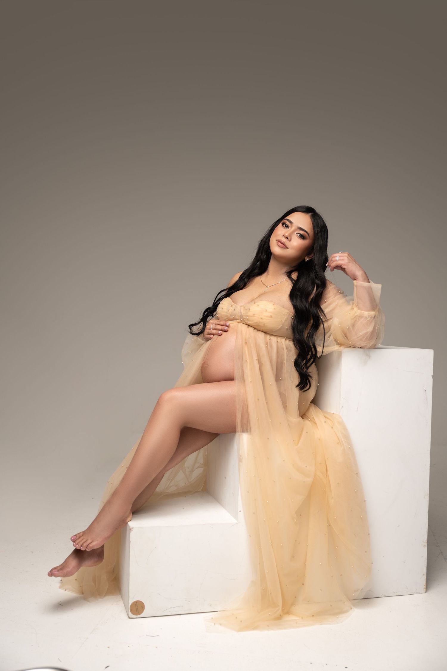 Pregnant woman in sheer golden robe poses gracefully on white blocks in a studio setting.