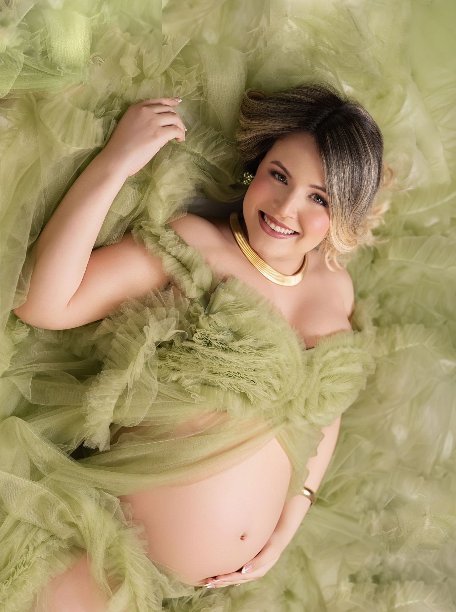 Smiling pregnant woman lying in green tulle fabric wearing a gold necklace, maternity photography session.
