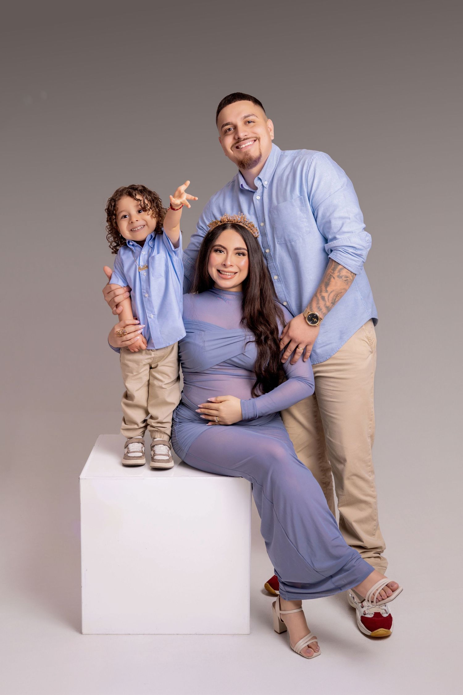Pregnant woman in blue dress sits with smiling husband and curly-haired son, all wearing coordinating blue outfits in studio.