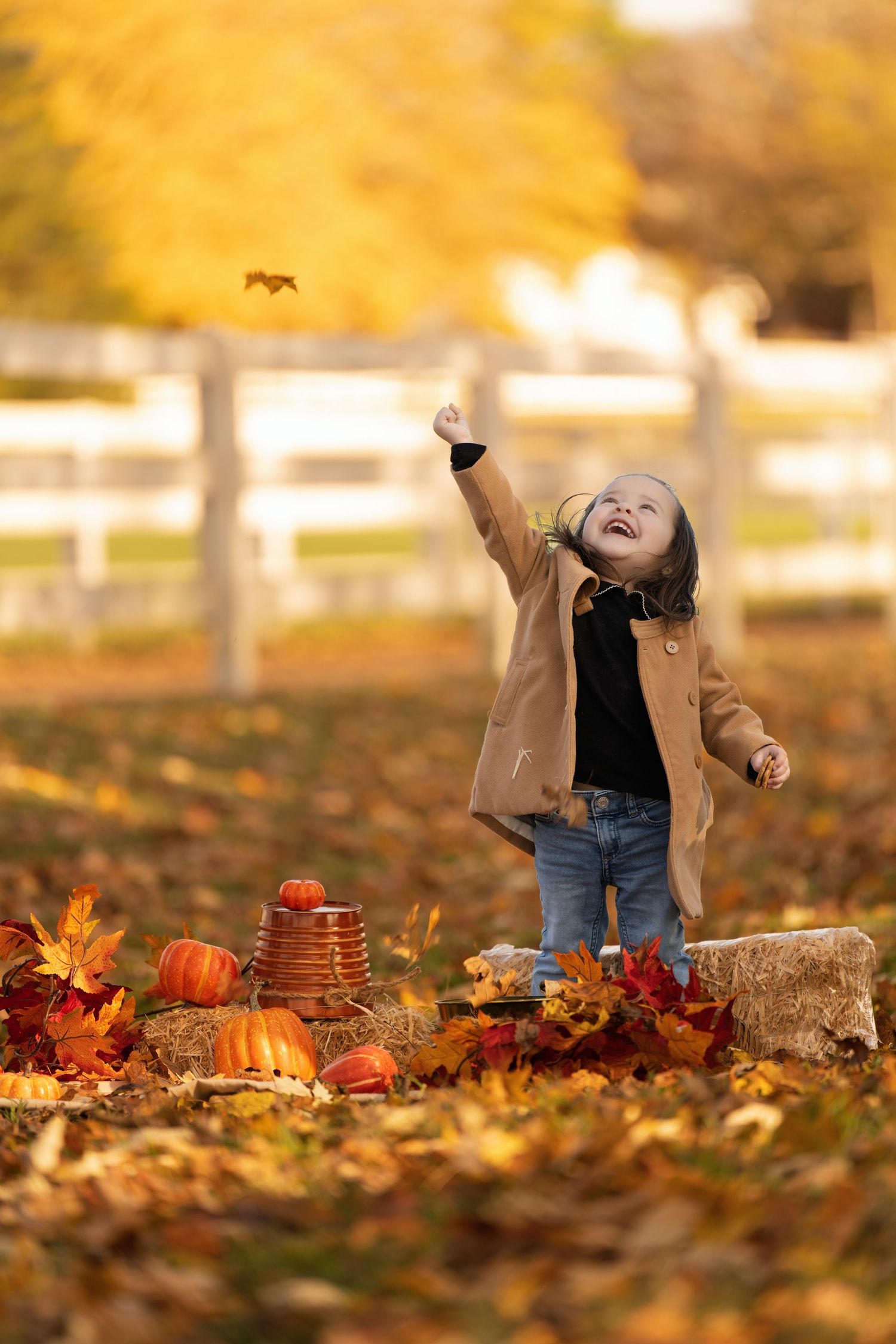 A joyful toddler in a tan coat tosses autumn leaves among pumpkins and hay bales in a golden fall outdoor setting.