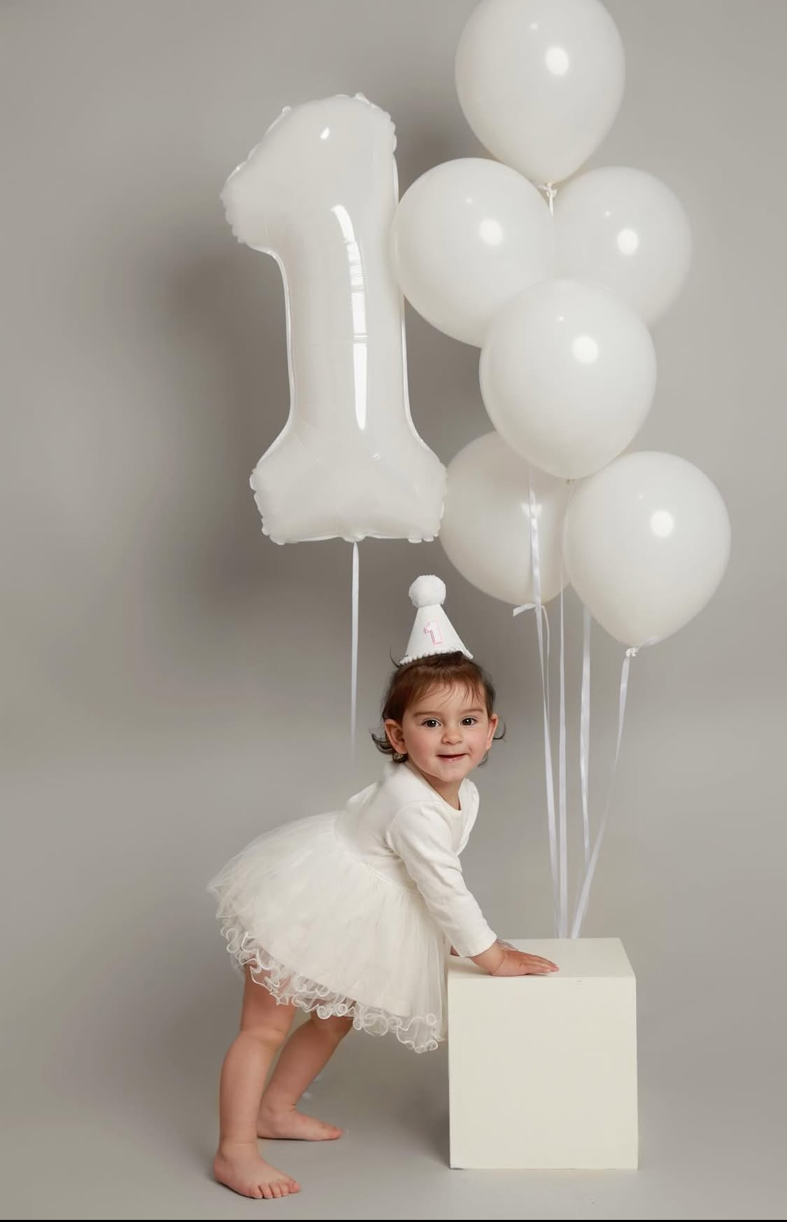 Smiling toddler in white dress and party hat poses by white balloons and number 1 balloon for first birthday.