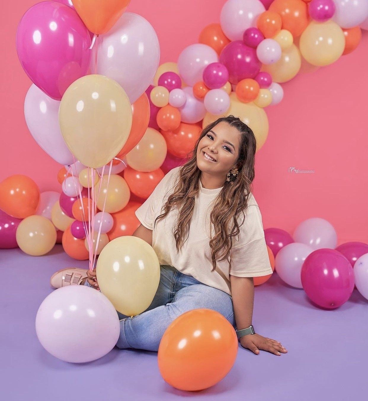 Young woman sitting on floor surrounded by colorful pink, orange, and cream balloons against a purple backdrop.