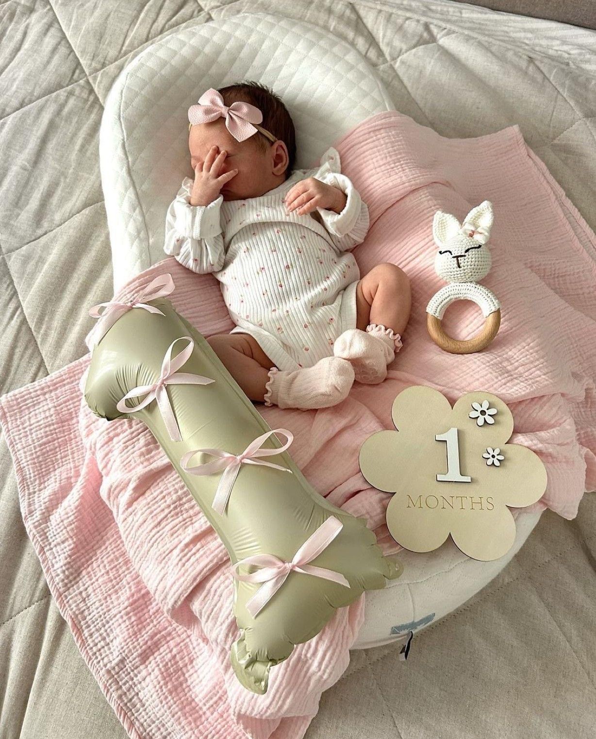 Newborn baby girl in white outfit with pink bow headband lying in snuggle nest with 1 month milestone sign.