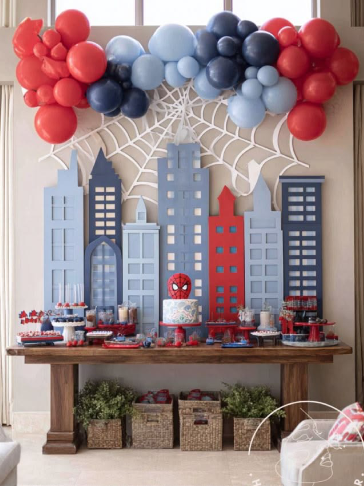 Spider-Man themed birthday party dessert table with cityscape backdrop, spiderweb decor, and red and blue balloon garland.
