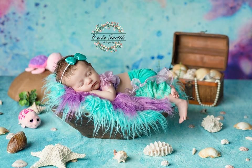 Newborn baby sleeping in a boat prop wearing mermaid costume with teal fur, shells, and treasure chest backdrop.