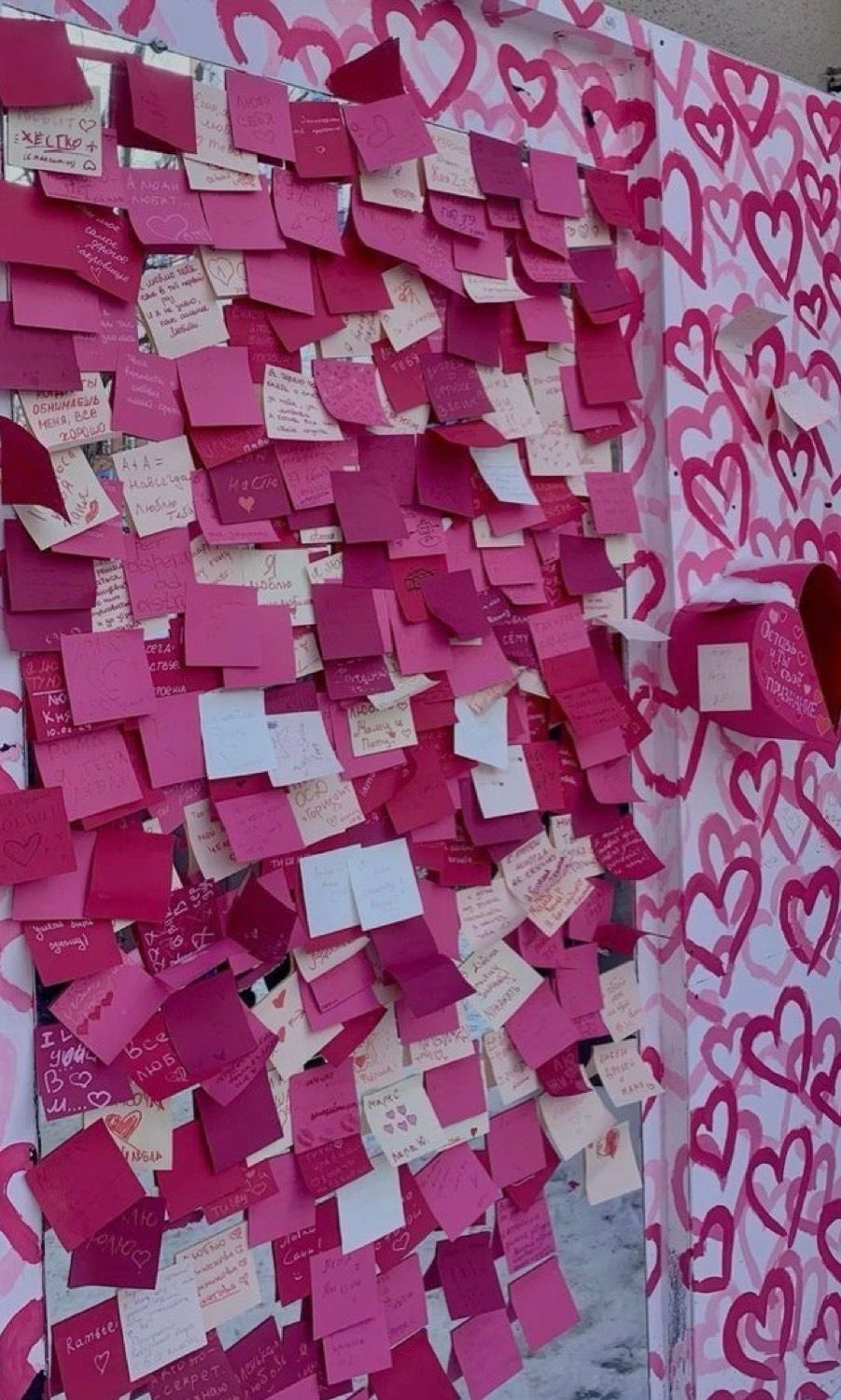A wall covered in pink and magenta sticky notes with handwritten messages, next to a heart-patterned pink mural.