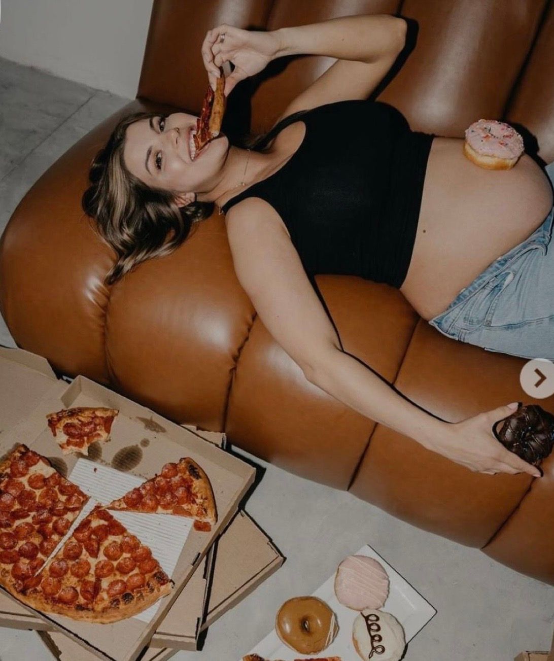 Pregnant woman relaxing on brown leather sofa eating pizza and donuts surrounded by food boxes.