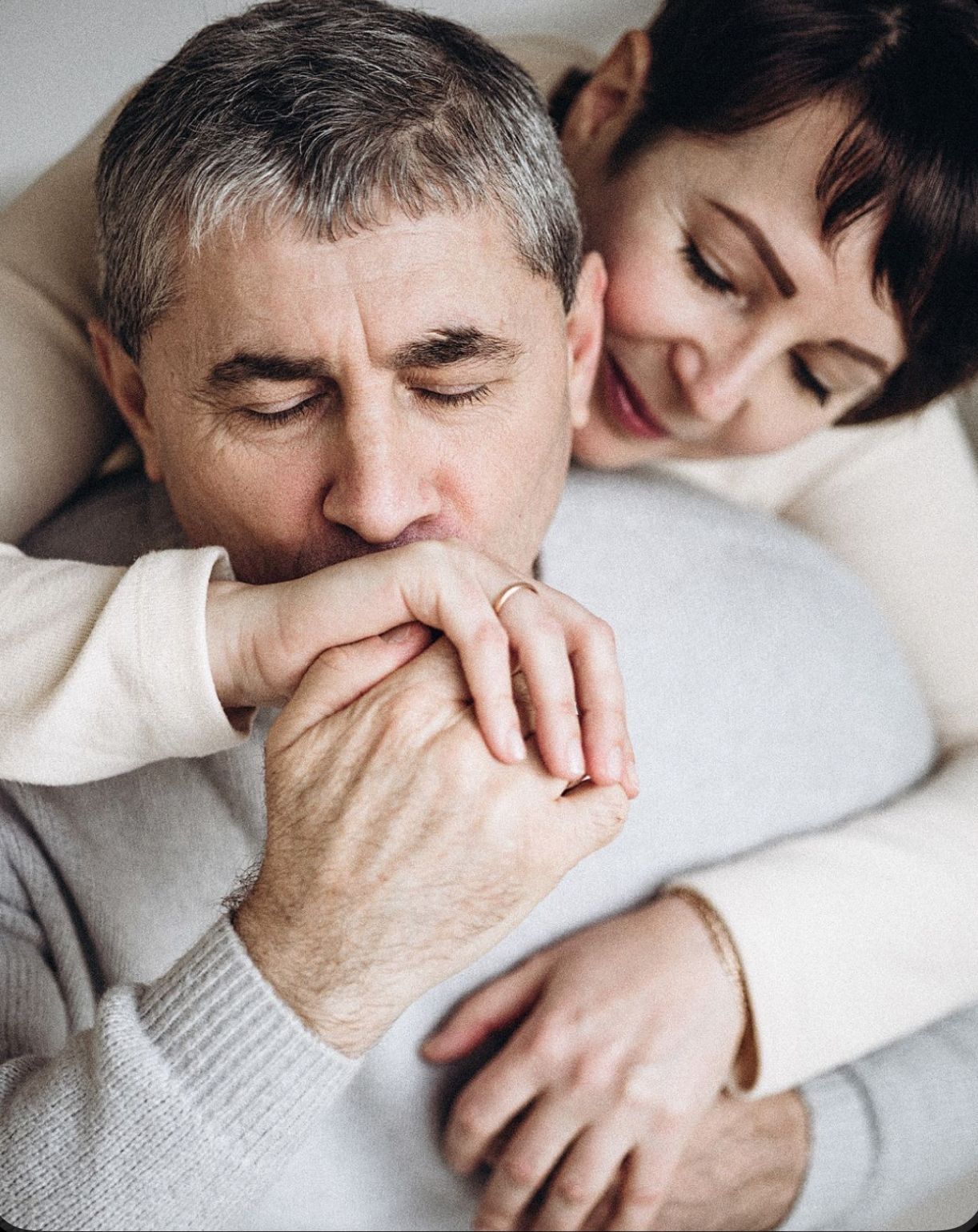 Middle-aged couple sharing a tender moment, man kissing woman's hands while she embraces him from behind.