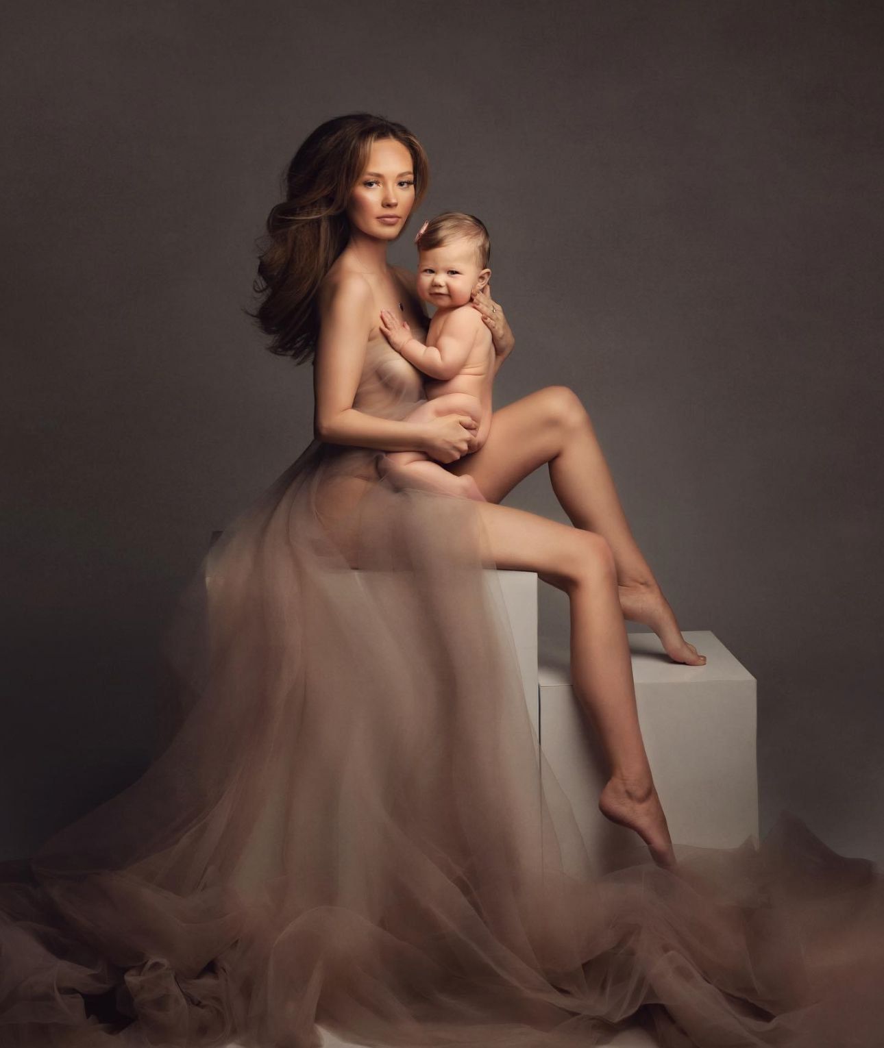 Mother in flowing tulle gown holds baby in elegant studio maternity portrait photography.