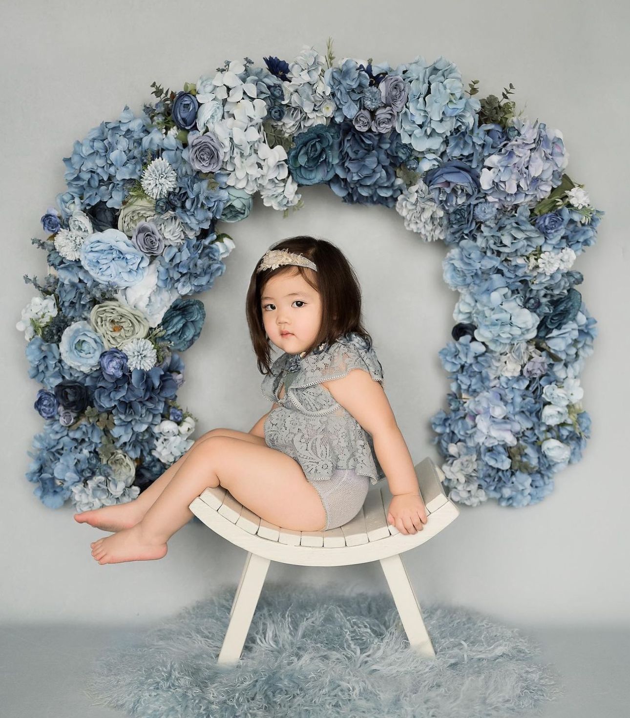 A child sits on a curved white bench against a beautiful blue and white floral arch backdrop in a studio setting.