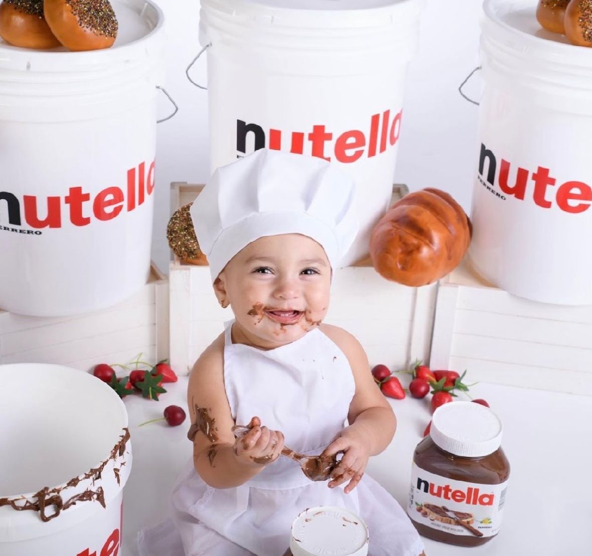 A small chef in white attire poses with Nutella jars and baked goods against a white backdrop.