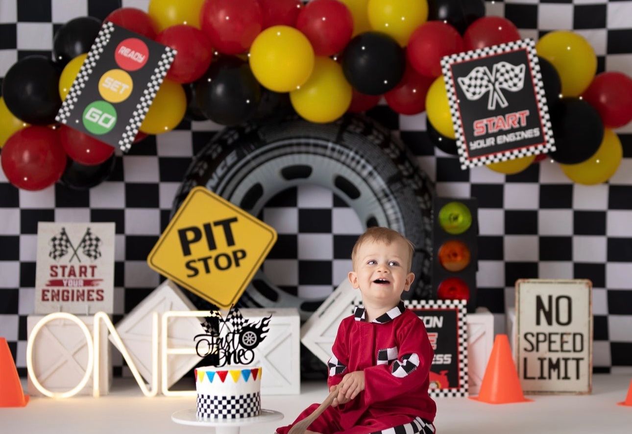 Racing themed birthday party setup with checkered flags, red, yellow and black balloons, and pit stop decorations against a gray wall.