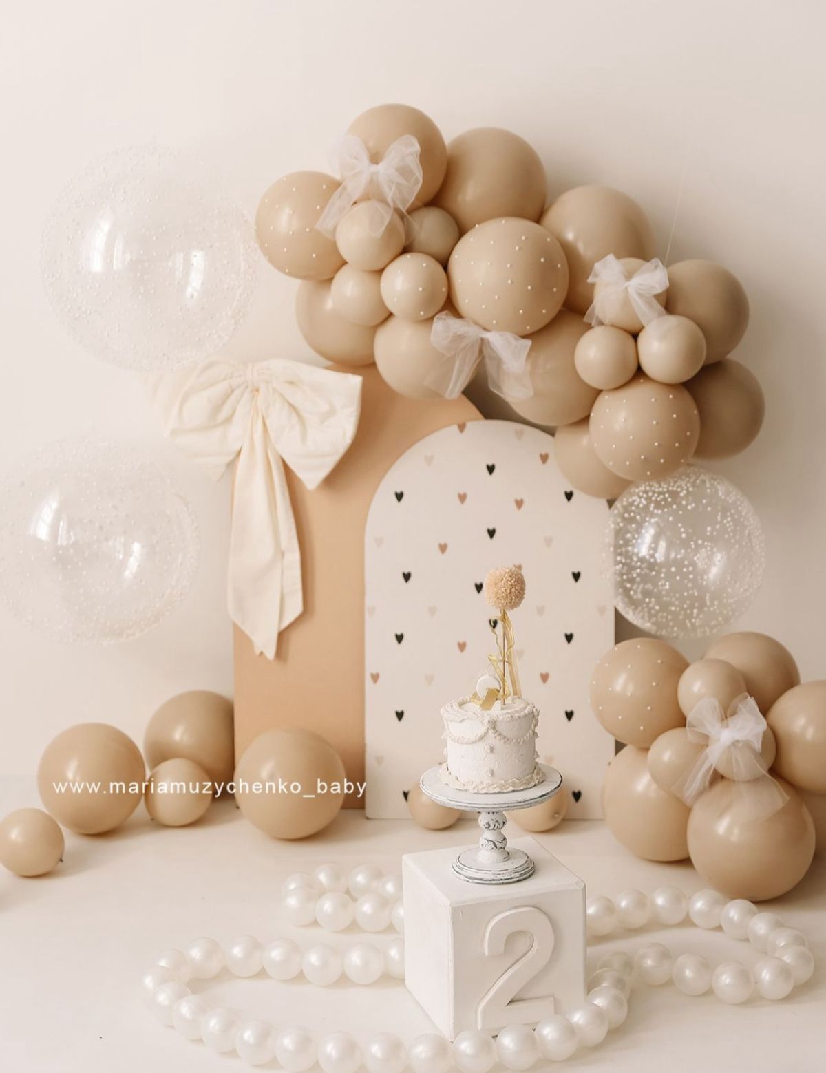 Neutral beige balloon arch with white bows, pearls, and small black heart decorations against a cream colored backdrop.