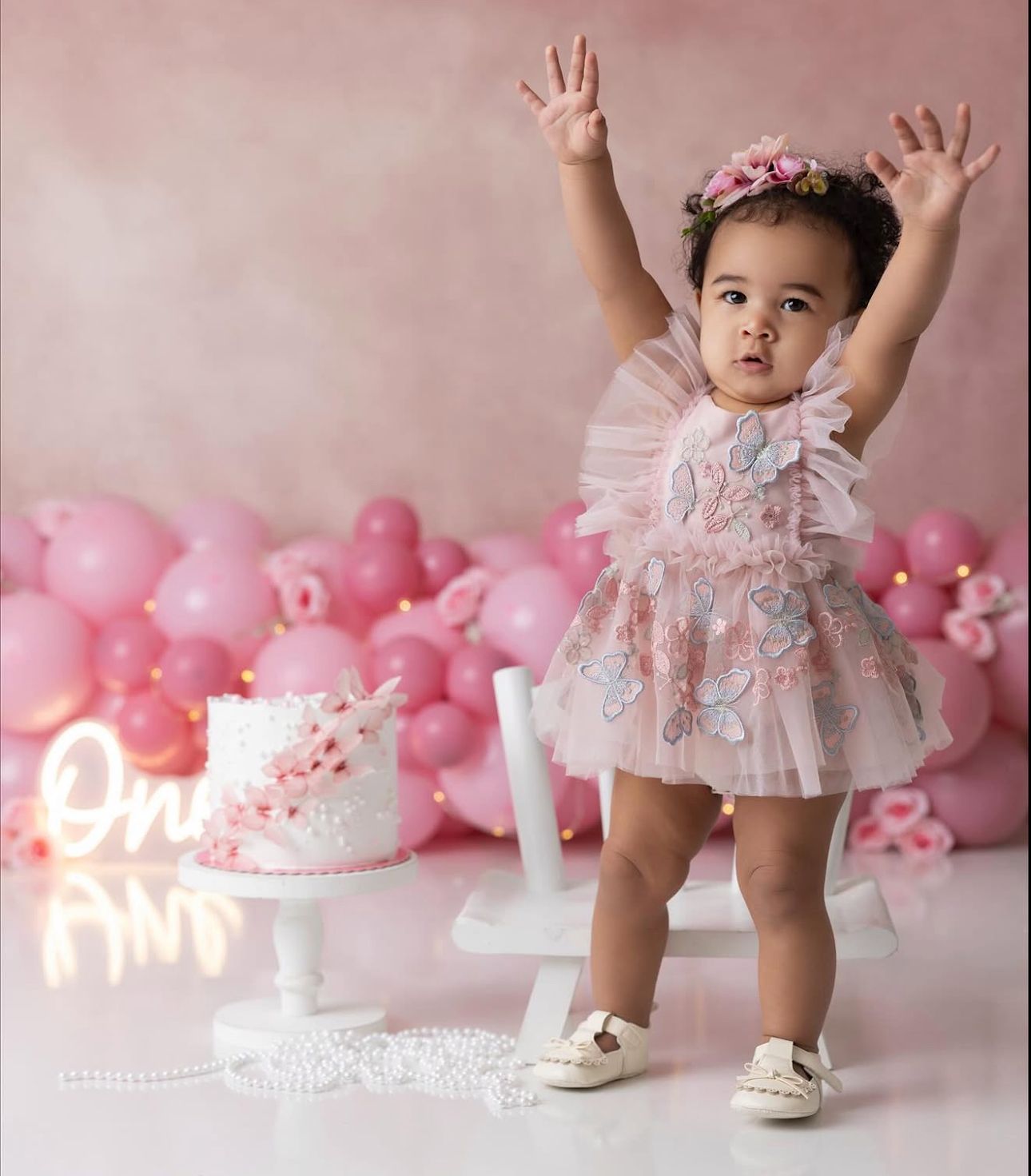 A small child in a lace dress poses with raised arms next to a birthday cake and pink balloon decorations.