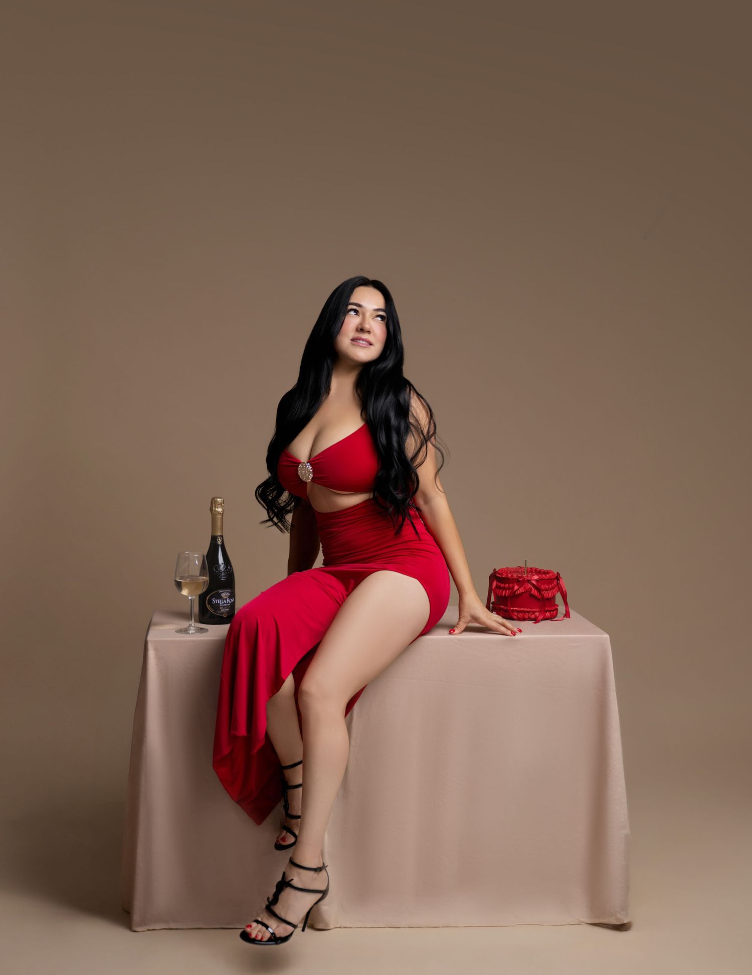 Woman in red two-piece outfit sitting on table with champagne bottle and red gift box on beige background.