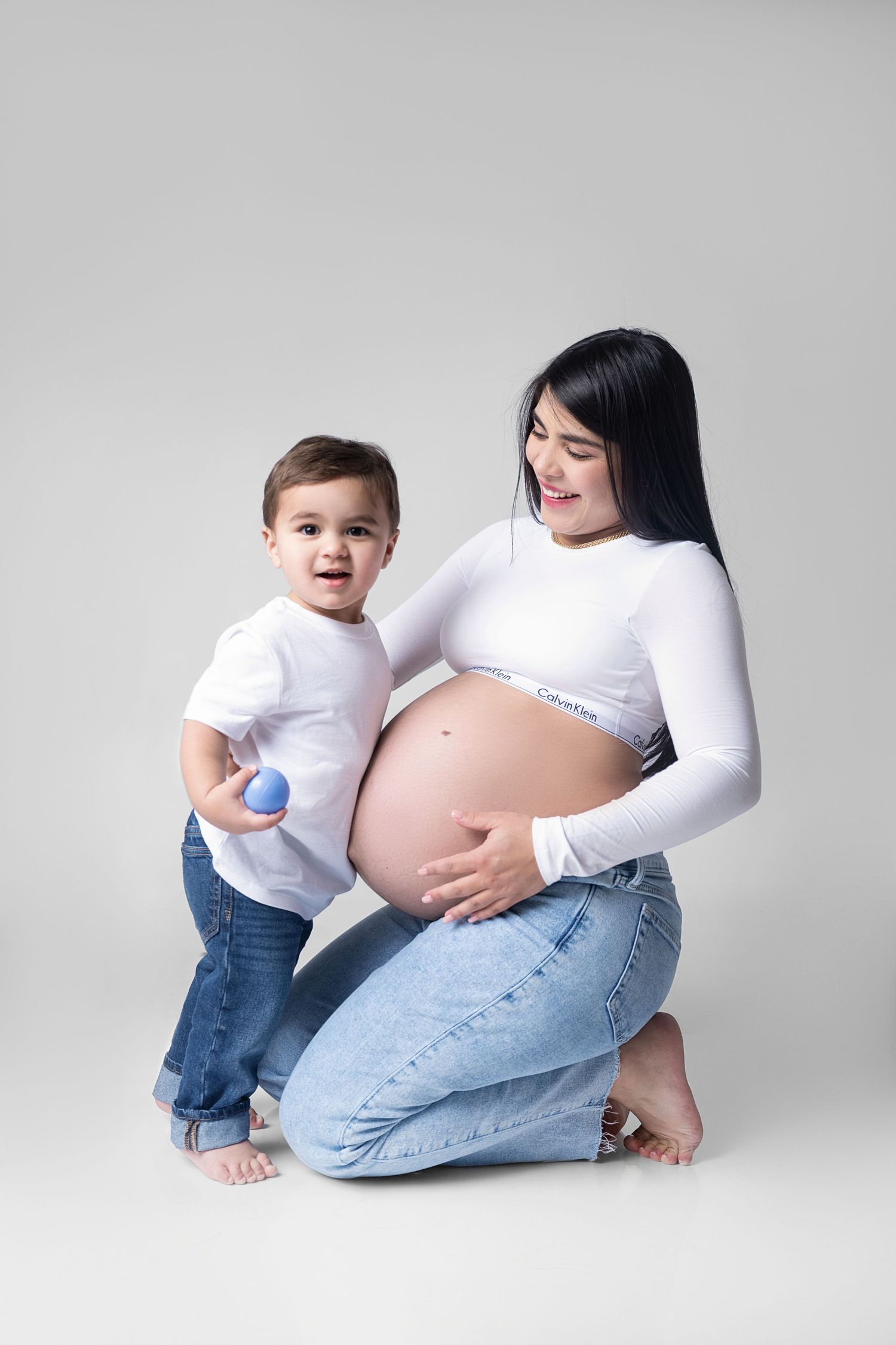 Maternity photo series showing tender moments between pregnant woman in white top and jeans with small child on white backdrop.