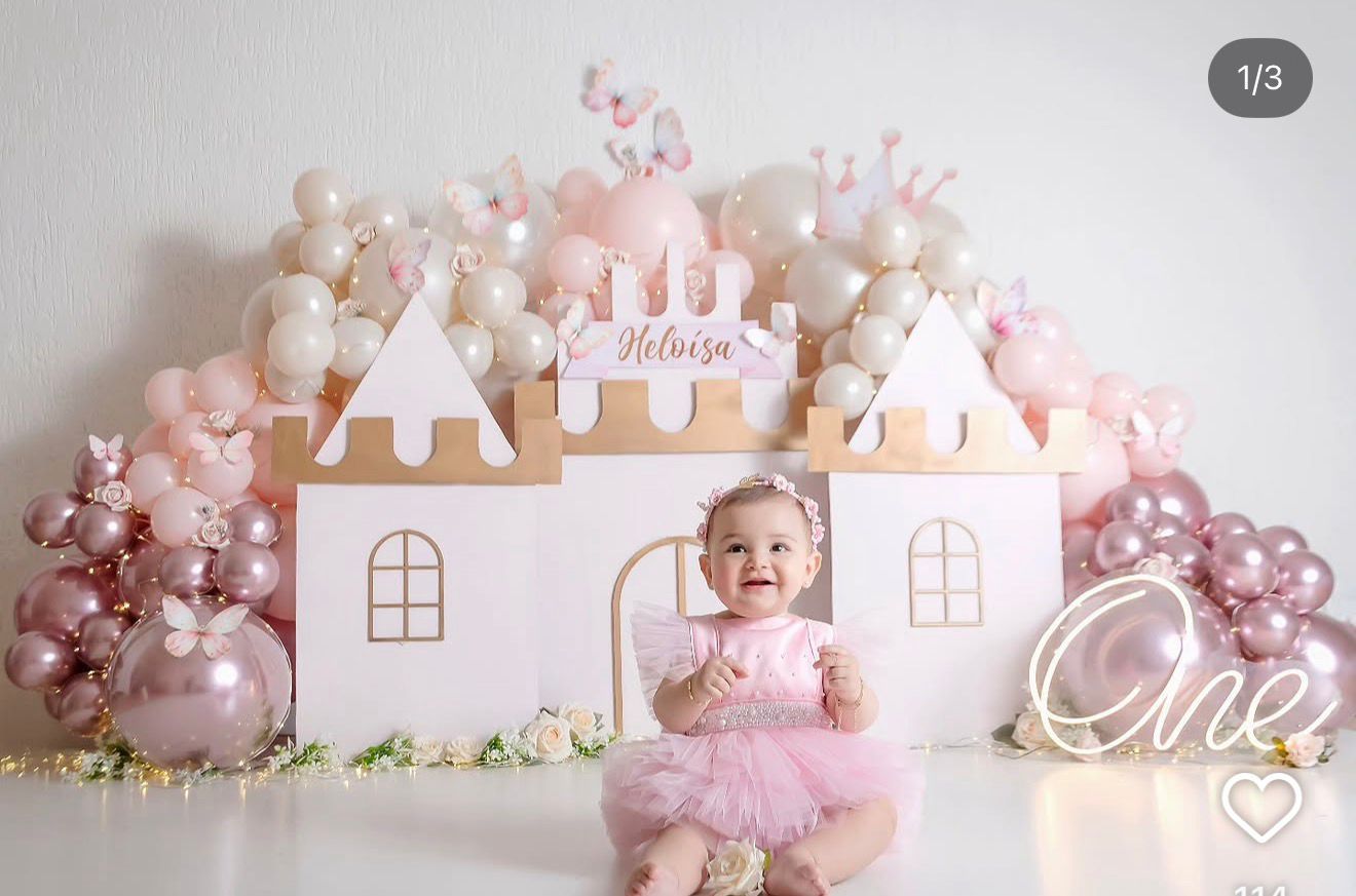 Baby girl in pink tutu sits before a castle backdrop with pink balloons and crown for a first birthday celebration.