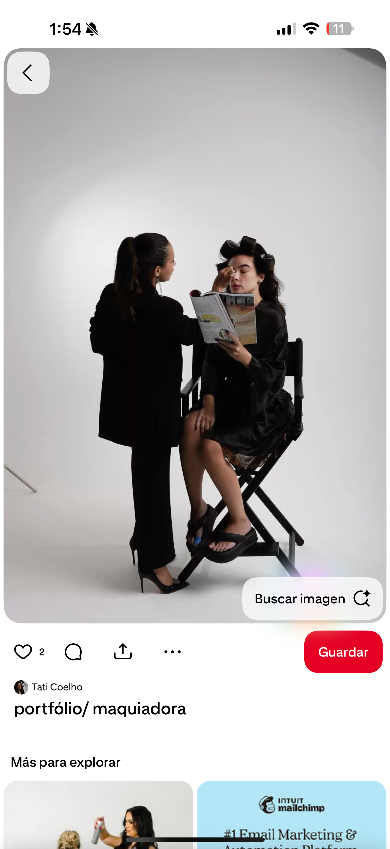 Makeup artist applying makeup to a model seated in a director's chair, both wearing black outfits in a studio.