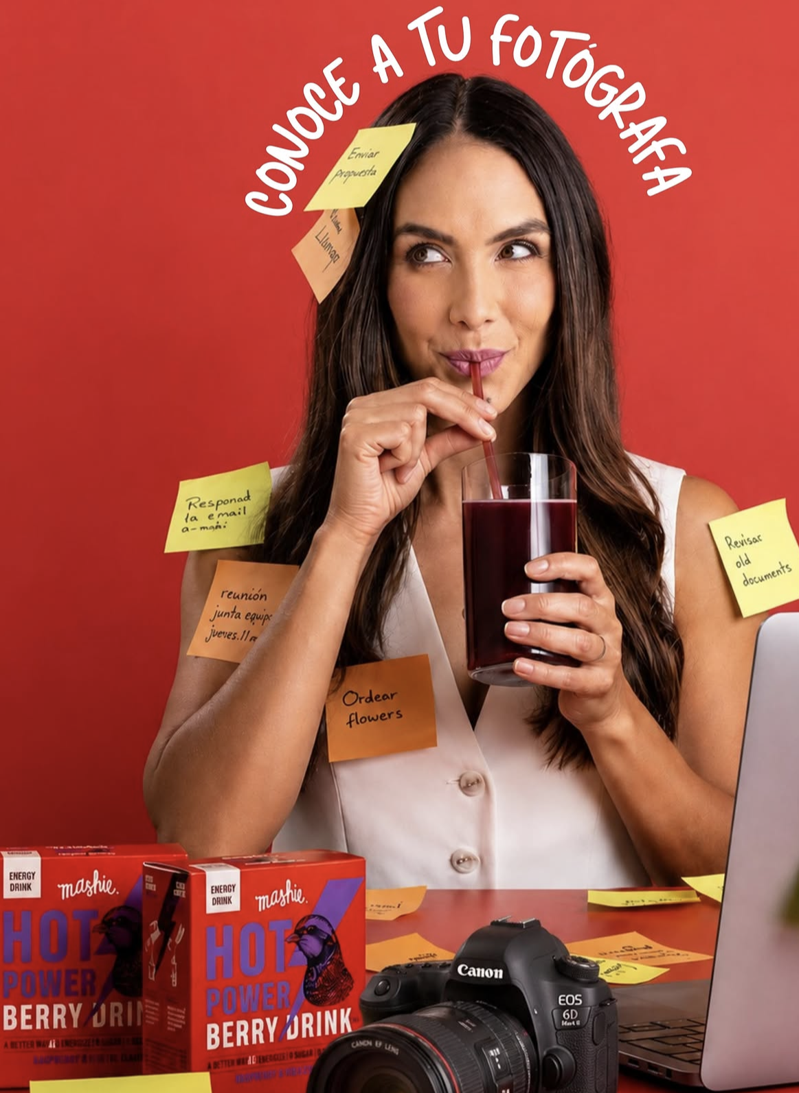 Woman sipping berry drink at desk with Canon camera, sticky notes, and Mahe Hot Berry Drink boxes on red background.