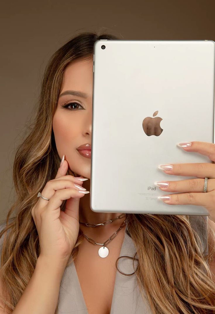 Woman with long highlighted hair holds a white Apple iPad in front of her face, wearing layered necklaces.