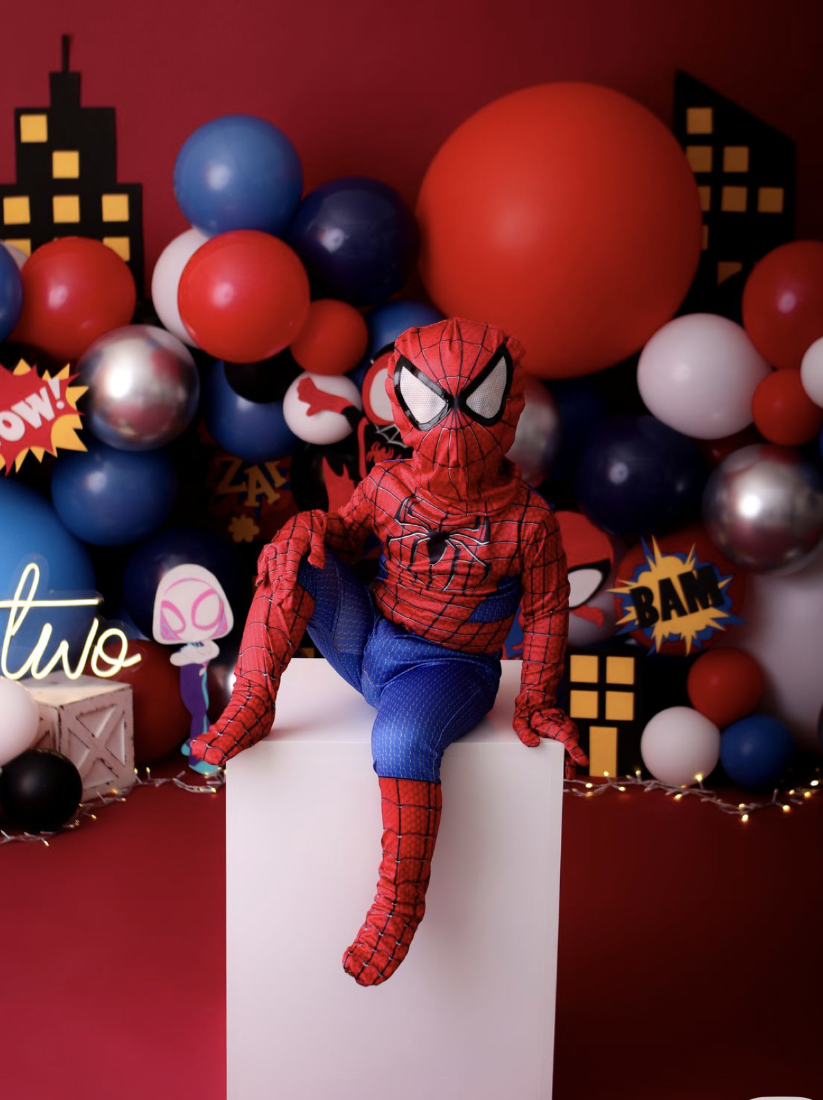 Child in Spider-Man costume sits on white block with colorful balloon backdrop and superhero birthday decorations.