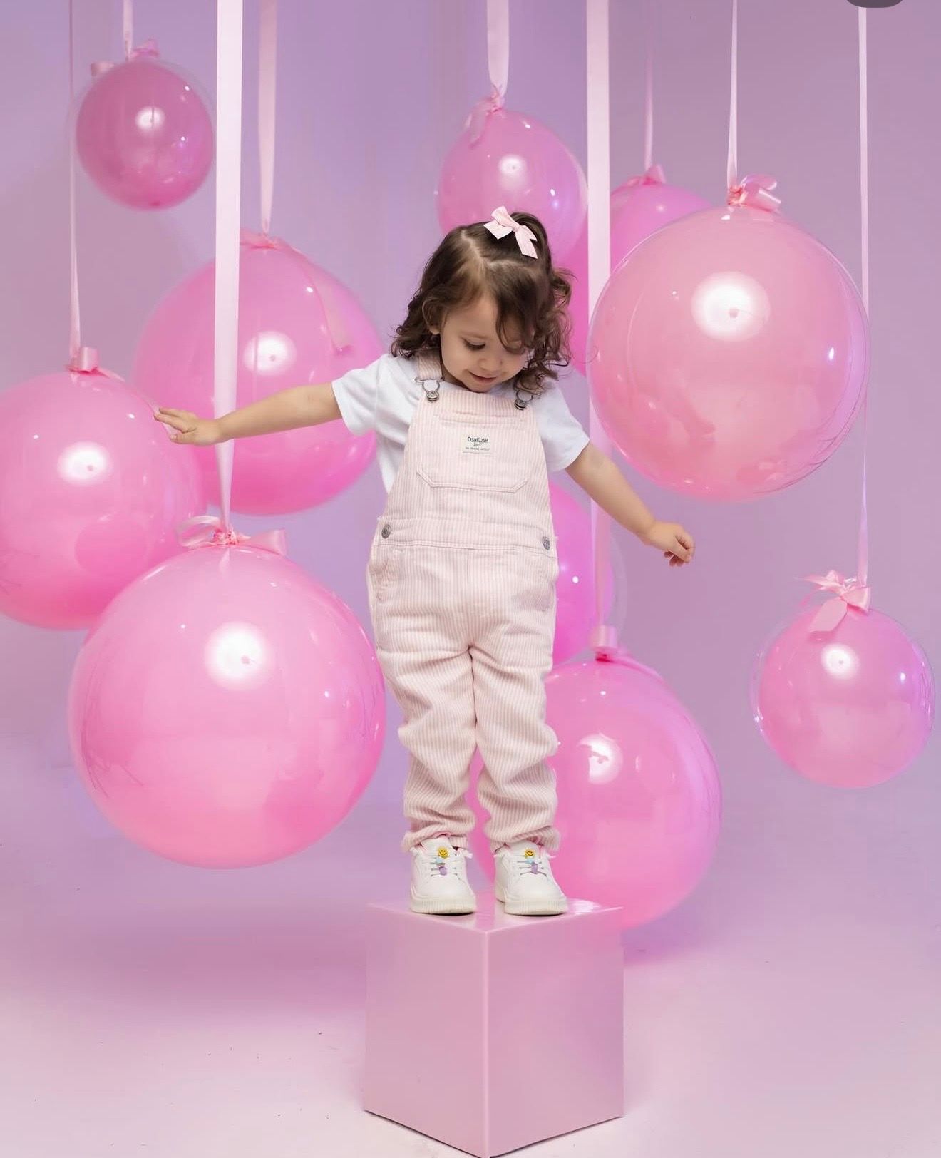 A toddler girl in pink overalls stands on a pink cube surrounded by hanging pink balloons in a festive pink setting.